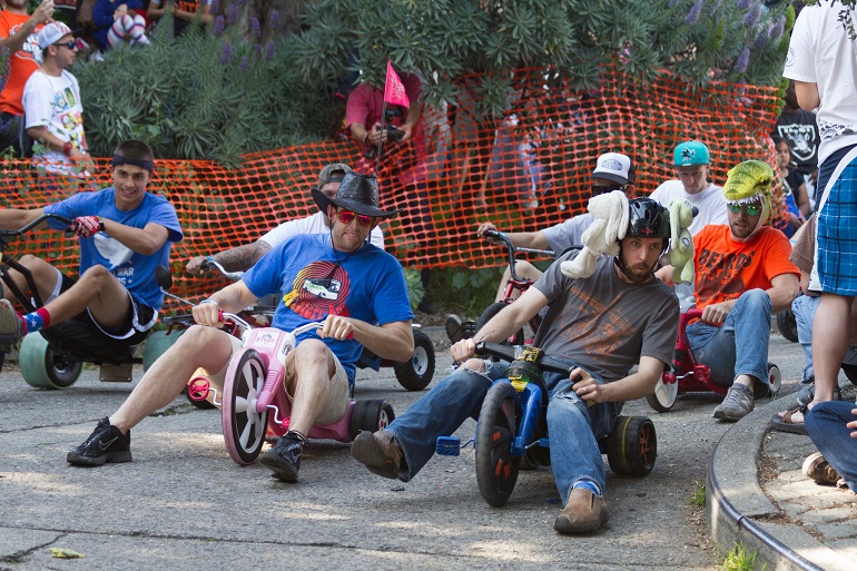 Men in brightly colored t-shirts and hats round the course of the Bring Your Own Big Wheel race with an orange mesh barrier behind them and spectators surrounding them.