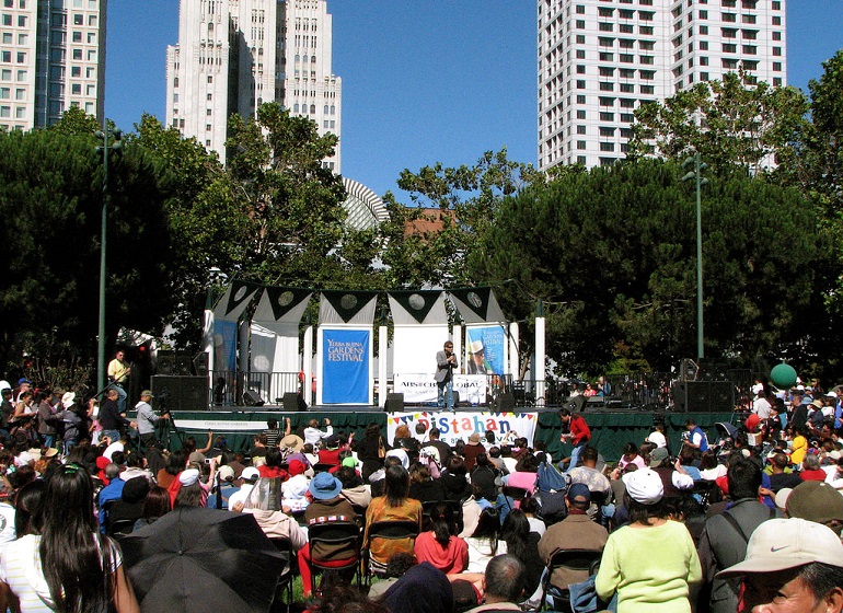 A crowd in a park watches entertainment on a small stage with skyscrapers and blue sky in the background.