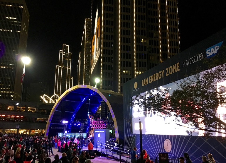 Crowd gathers at night in front of the concert stage at Super Bowl City with downtown skyscrapers in the background.