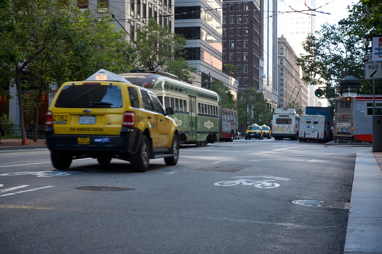 A  yellow taxi, a historic streetcar and Muni buses crowd their way down a busy Market Street.