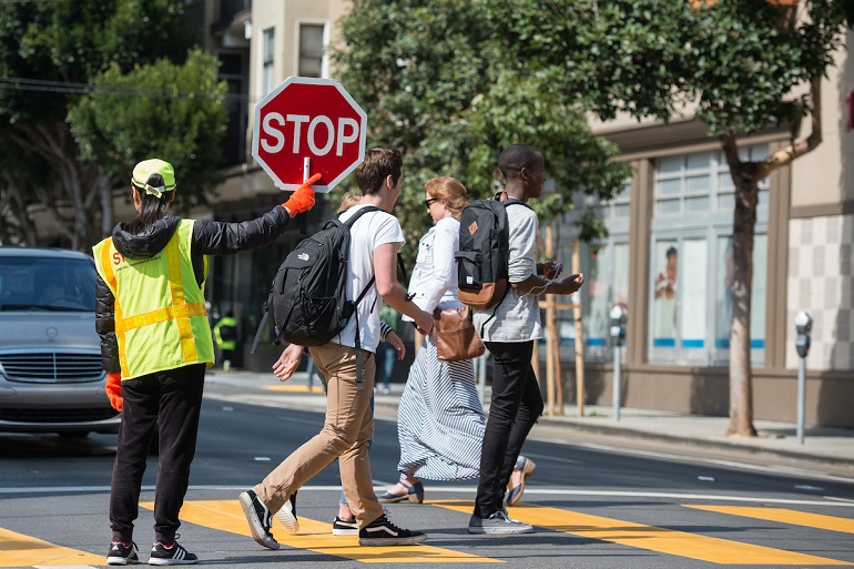 A pedestrian in yellow reflective gear holds up a stop sign while pedestrians with backpacks cross in front of her.