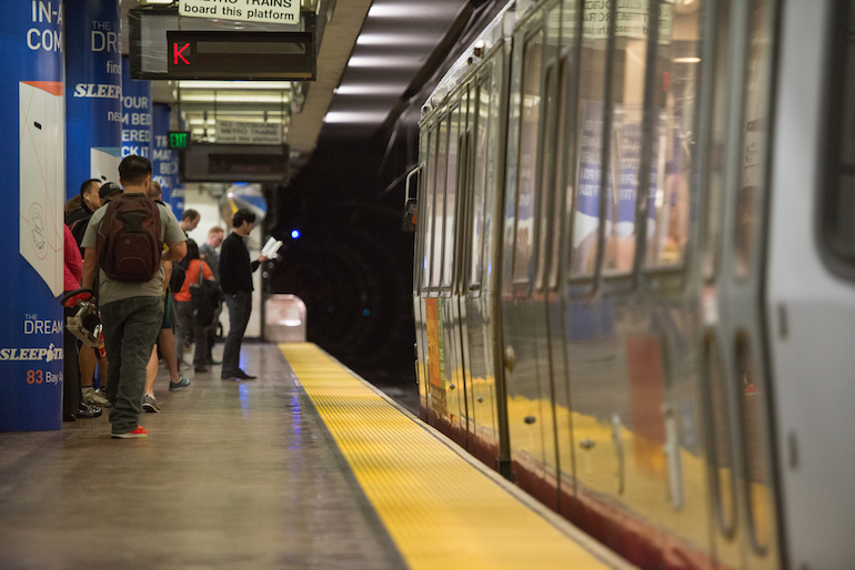 Muni customers stand behind the bright yellow strip on a subway platform as the light rail train pulls in on the right.