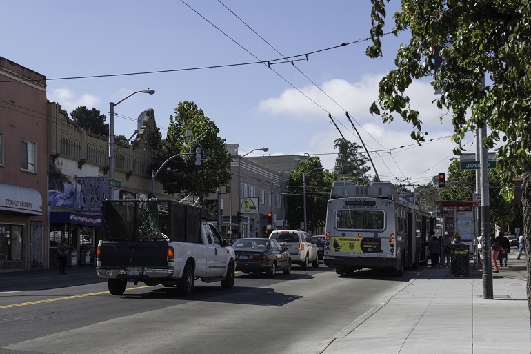 Muni trolley bus sits at a shelter stop while passengers board. A column of cars and trucks wait at the red light next to it.