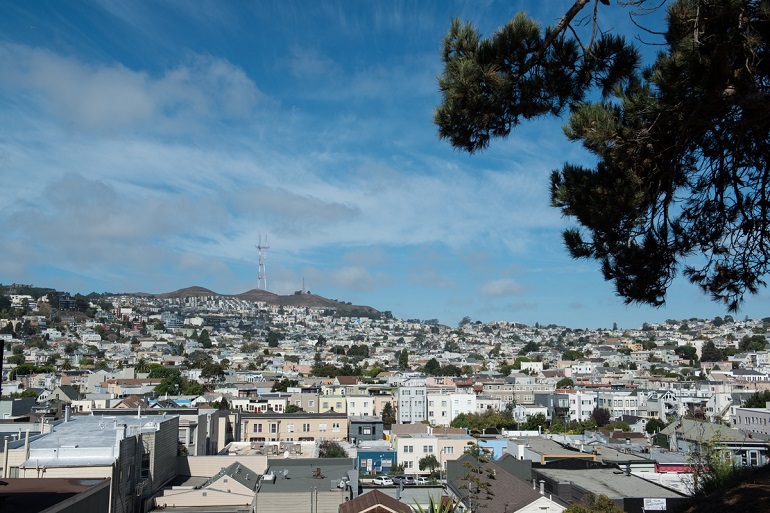 View from Bernal Heights of Mission District rooftops, Sutro Tower and a blue sky with scattered clouds.
