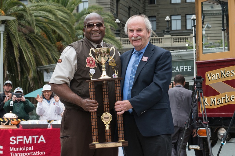 Two men, one in a suit and the other in a brown Muni operator's uniform, stand together holding a large trophy with a motorized cable car in the background.