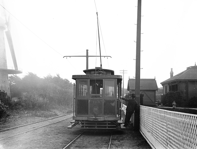 Circa October of 1903, a man hangs off the side of an old-fashioned streetcar on an unpaved trackway in the outskirts of Golden Gate Park. To the right, a low wooden fence separates the trackway from two small house-like buildings that served as the U.S. Life-Saving Station. To the left is a partial view of trees, shrubbery and the “Dutch” Windmill.