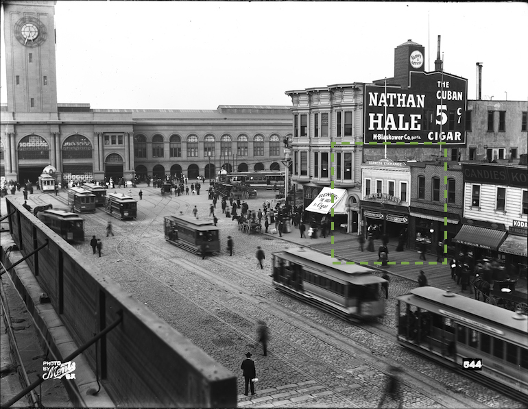 Black and white photograph taken August 22, 1905 showing lower Market Street between Steuart Street and present day Embarcadero (then called East Street). In view are the Ferry Building and cable cars. Across Market Street is a row of buildings and storefronts, below which lies a wooden plank sidewalk with people walking. One building in the image is highlighted by a box drawn in a green dotted line on the image to indicate the area detailed in the next image.