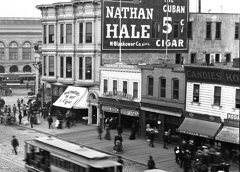 A zoomed-in view of an area highlighted in the previous photograph, showing a view of Market Street near today's Embarcadero, showing a busy wooden sidewalk and businesses with Victorian storefronts, including the Alameda Exchange.