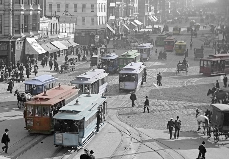 An image from the SFMTA Photo Archive from 1905 looking west on Market Street with a variety of cable cars in shades of green, blue, off-white, red and yellow. The colors in this image have been added by hand and the background of the image is still in shades of grey. There are many pedestrians on Market Street as well as horse-drawn vehicles.