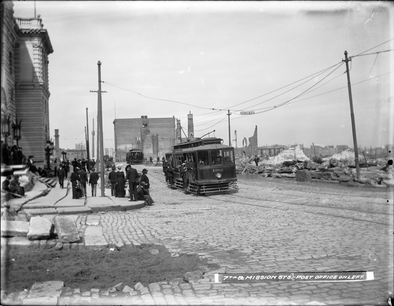 view east on Mission and 7th streets of earthquake destruction, buckled cobblestone street and electric streetcar full of people riding past US Post Office building.  May 9, 1906