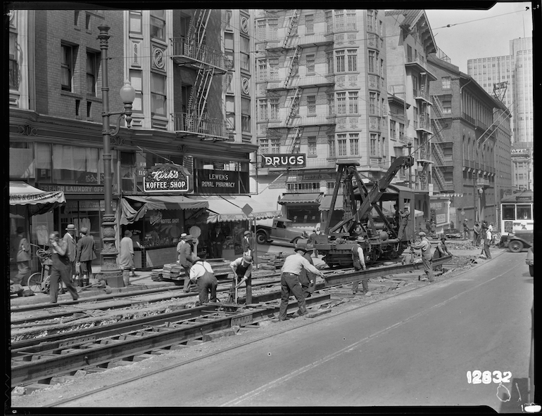 A black and white photo taken in 1931 shows a wide view of the intersection of Sutter and Jones streets,, looking northeast on Sutter towards downtown. In the center is a group of men laying streetcars rails on an open trench. Behind them and further down Sutter is a flat railcar equipped with a crane, which is lowering a long section of rail onto exposed railroad ties. Beyond that is a streetcar with the front painted white, operated by the Market Street Railway Company. Along the sidewalk behind the men working in the street is a row of storefron businesses including a "French Laundry" "Kirk's Koffee Shop" and, on the corner "Lewin's Royal Pharmacy", a drug store.  Some pedestrians stand nearby.