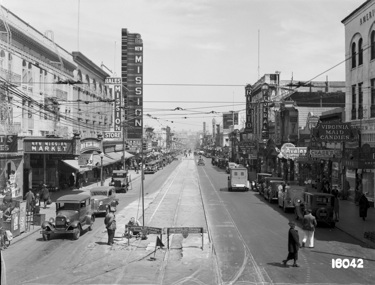 Overhead view looking north on Mission Street from 22nd street showing new streetcar tracks.  Theatre marquees, storefronts, pedestrians, and autos along corridor from foreground to background.  July 24, 1936