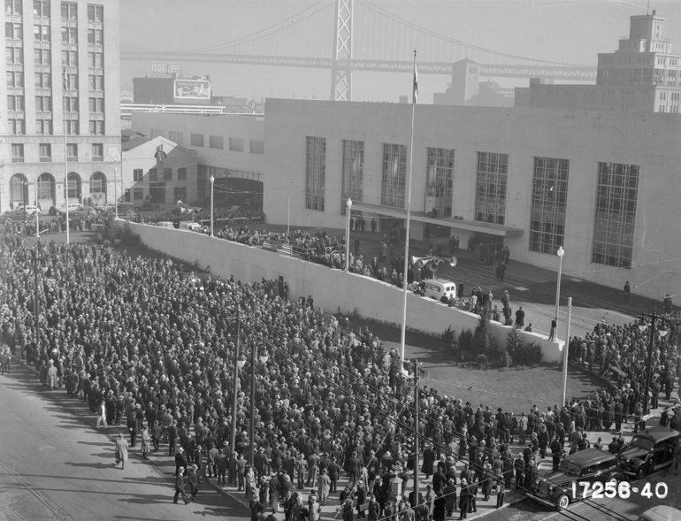 Overhead view of crowd of thousands of people gathered for the opening of the original Transbay Terminal on January 14, 1939 with San FRancisco-Oakland Bay Bridge and downtown buildings in background.