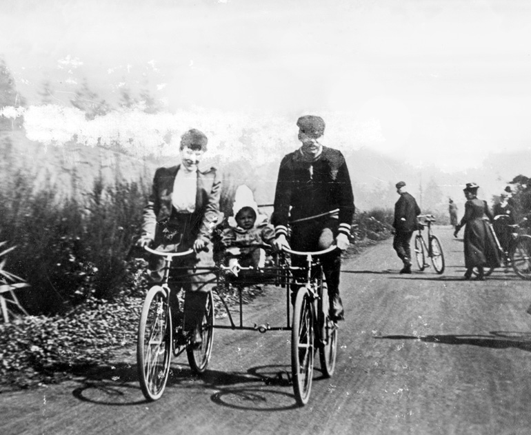 A black and white photograph of a woman and a man riding two bicycles that are joined in the middle to support a seat for a baby. The people in the image wear period garb of the 1890s and are cycling through Golden Gate Park.