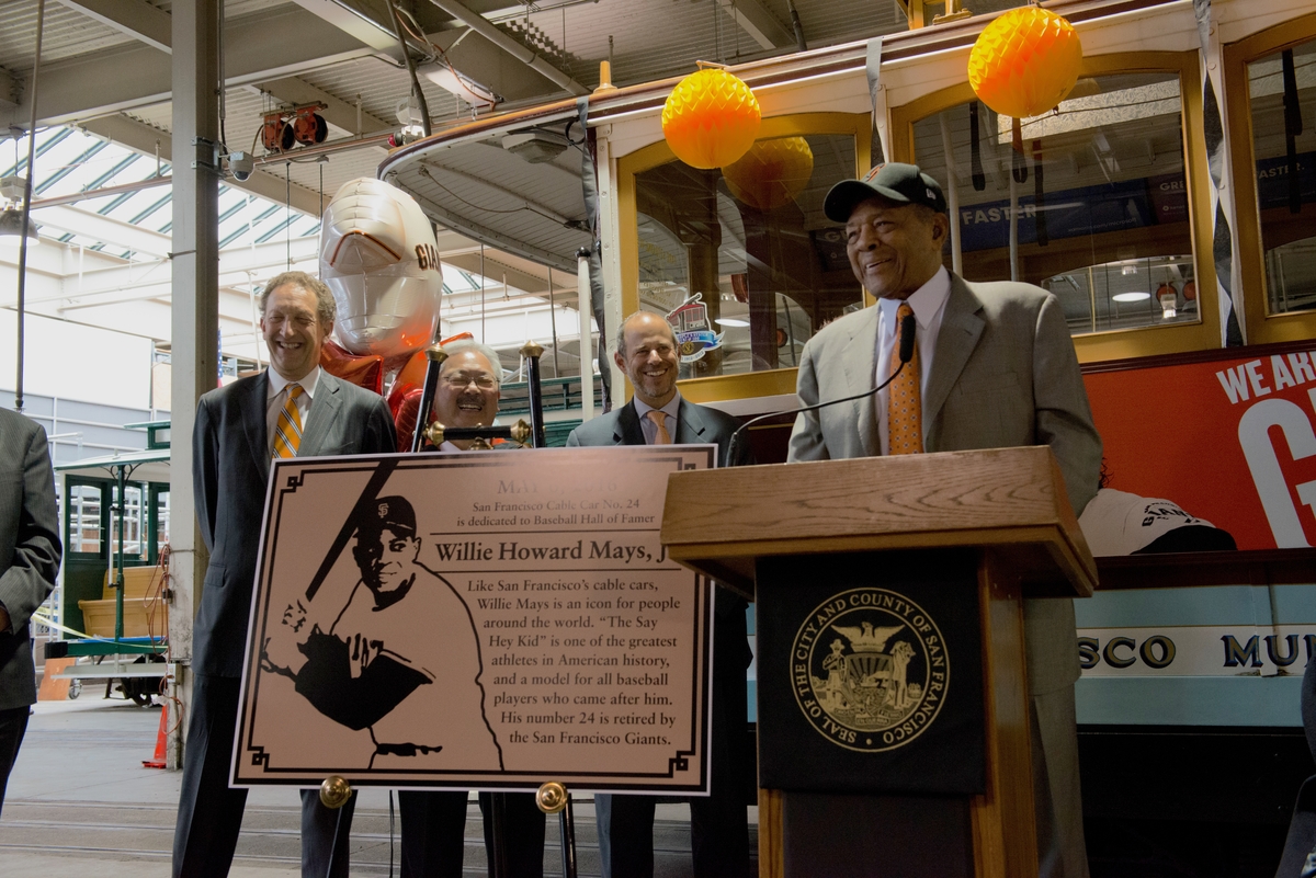 Four men stand behind a podium and a replica of the Willie Mays plaque. A cable car decorated with balloons and streamers sits behind them.