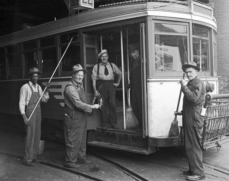 Men in coveralls with brooms sweep up an old-fashioned streetcar inside of a streetcar barn. The streetcar is painted with a stylized representation of wings on the side and has a 22 sign for the 22 Fillmore route. The image is in black and white and was taken in 1948.