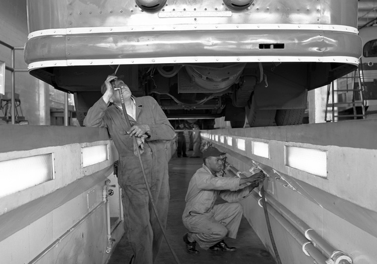 A shiny 1940s bus is parked above a below-ground pit in which two men are working on the underside of the bus. Inside the pit is clean and is light by bright panels set into the sides of the wall in a geometric, modern style. The image is in black and white and was taken in 1949.