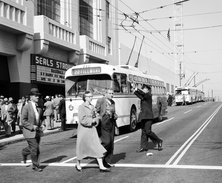 People in trench coats and fedoras walk in a crosswalk in front of a bus. The bus is dropping off passengers for a baseball game at Seals Stadium, located at 16th Street and Bryant Street. The image is in black and white and was taken in 1958.
