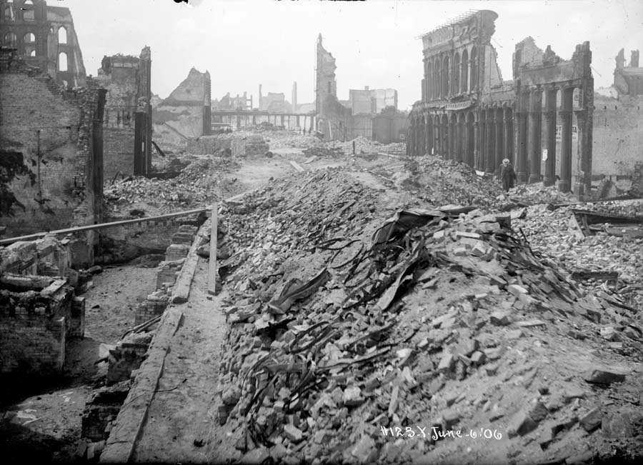 Black and white photograph showing burned and crumbled buildings after 1906 earthquake.  Taken at California and Battery streets on June 6, 1906.  In the foreground are large piles of rubble filled with broken bricks, metal and wire scraps and in the foreground are partially collapsed skeletal building walls standing with no windows or interior floors and walls.