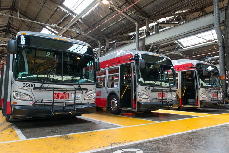 color photo taken in 2016 inside a Muni bus garage showing the fronts of three buses lined up in a row.  These buses are grey with red stripes along the top and bottom of the bodies.