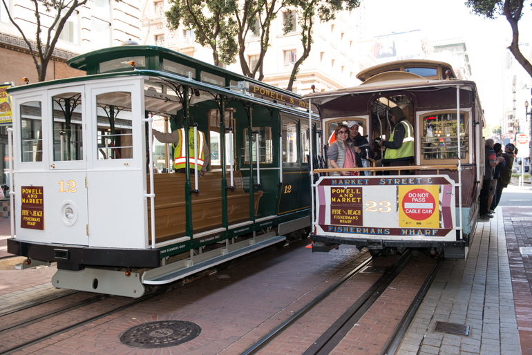 color 3/4 view of two cable cars, 12 and 23 on Powell street near Market.  Cars are passing each other on separate tracks, one is empty and the other is filled with passengers.