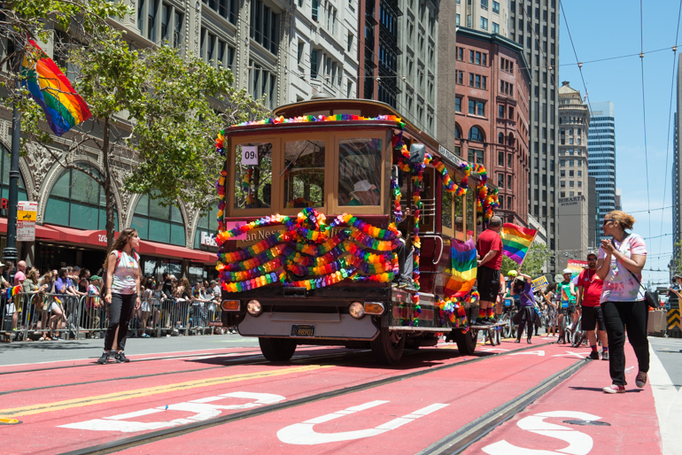 Color photograph from June 26, 2016 showing Muni motorized cable car 62 decorated in rainbow banners on Market Street as part of Pride parade.