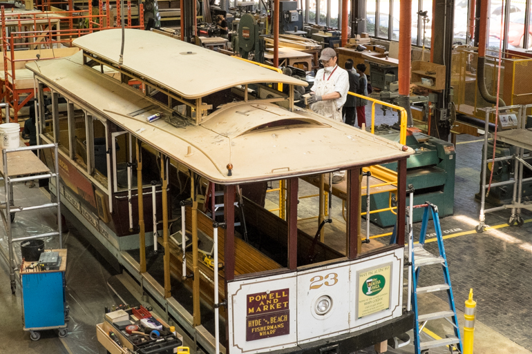 Overhead view of Cable Car 23 inside the carpentry shop.  A man is standing on a ladder next to the car while removing a part from the roof of the car.