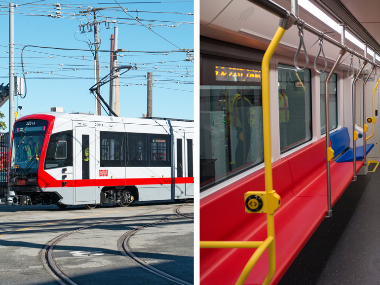 On the left side of the image, a new red and silver train with a large window on the front is parked in a San Francisco train yard on a sunny day. On the right side of the image, the inside of the train can be seen with red longitudinal setting and bright yellow stanchions.