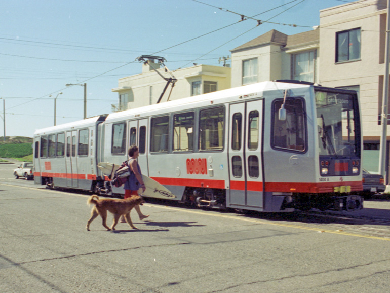 One of the current style of Muni trains, a boxy grey vehicle with a red stripe and the Muni worm logo, waits for passengers to board on Judah Street near Ocean Beach. Sand dunes are visible in the background, and a barefoot man with a surf board and a shaggy dog is approaching the train.