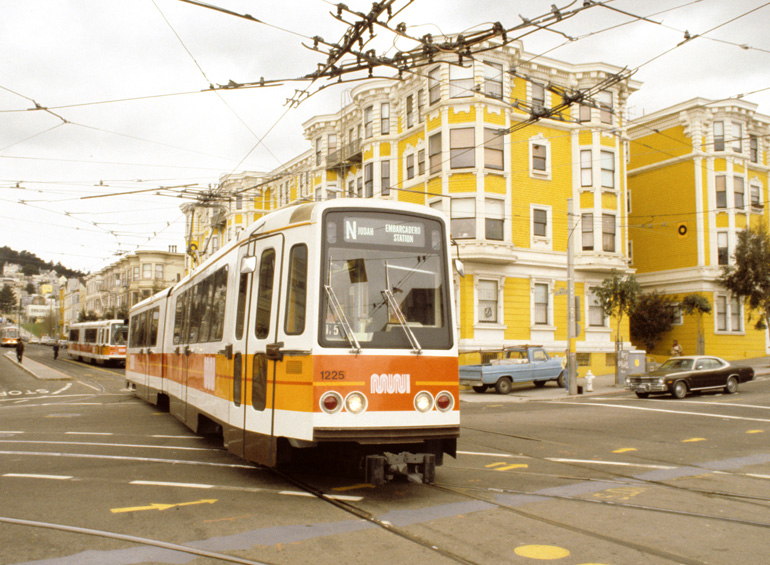 A mid-century style Muni light rail vehicle with orange stripes and the Muni worm logo is crossing the intersection at Church Street and Duboce Avenue. In the background a bright yellow building and late 1970s style vehicles are visible.