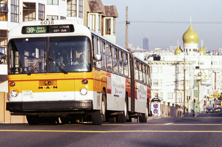 color photo taken in the 1980s looking east from Geary and 31st showing a long Muni bus painted in the "sunset livery", white with an orange and yellow stripe around the center of the bus.  In the background the golden onion shaped domes of a church rise above the cityscape.