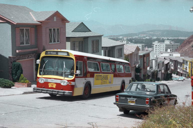 color photo showing a red, yellow, and silver painted Muni GMC bus climbing a hill in the Twin Peaks neighborhood in 1969.  Agreen volvo sedan is a right and the golden gate bridge towers can be seen faintly in the background.