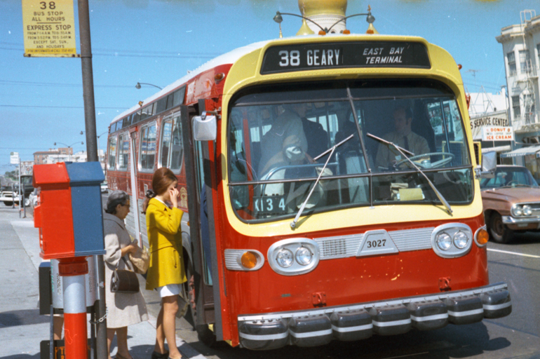 color photo taken in 1969 showing a red, yellow, and silver painted muni bus stopped at a stop on Geary Boulevard.  Two people, including a young woman wearing a mustard-yellow overcoat wait to board the bus.