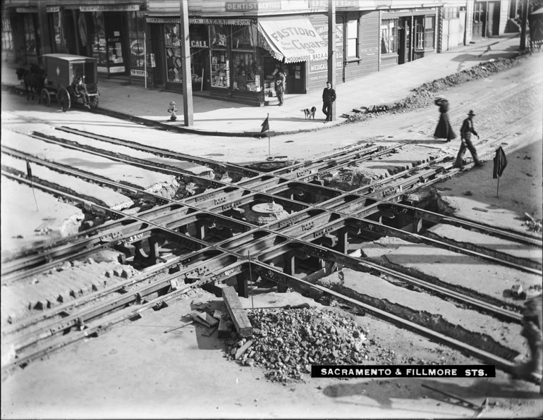 black and white photo from 1905 of intersection of Fillmore and Sacramento streets.  Track construction is taking place in the intersection while people walk around the crossings.