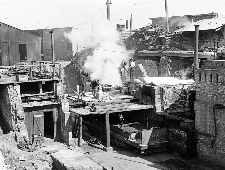 Black and white photo taken in 1910 of men working in an asphalt plant. A rail car sits in a brick-walled pit, above which men are moving steaming bins filled with hot asphalt.