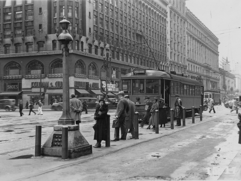 Black and white photo showing Market Street at Stockton on April 15, 1935.  In the foreground is a streetcar boarding platform with passengers standing in wait.