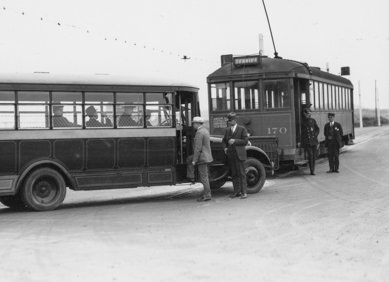 black and white photo showing people boarding a Muni bus in 1925 at the end of the L Taraval Line.  An L Taraval streetcar is seen in the background, amidst a wide open area of sand dunes in the undeveloped Sunset District.