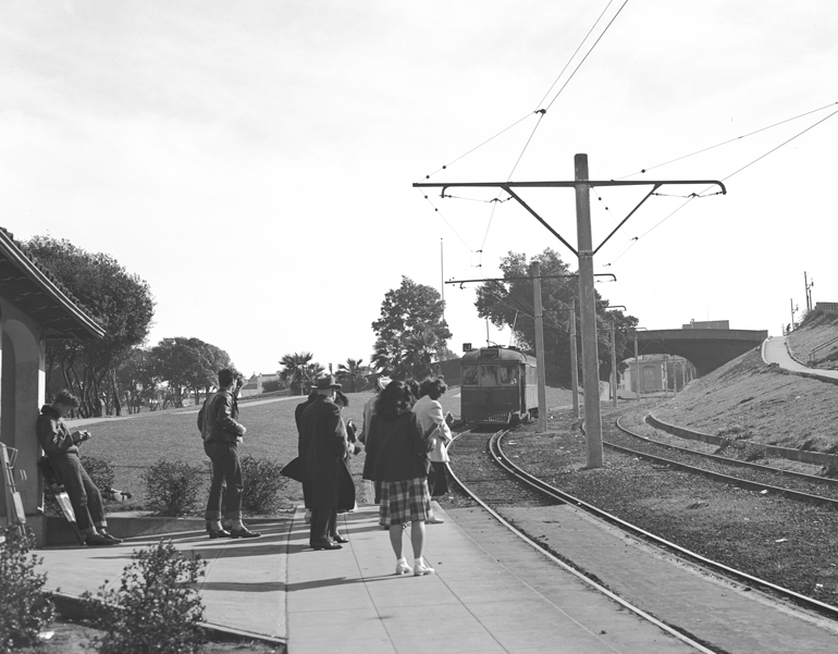 Black and white photo taken in the 1940s looking south from 18th and Church streets. Students wearing '40s era clothing wait as a J Church streetcar comes down the hill in Dolores Park.