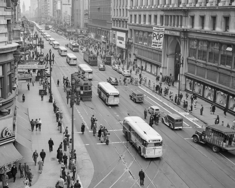 A black and white photo taken in 1948 looking east on Market Street from Powell, showing a long line of white colored motor buses parading down the street.  Taken from overhead, people stand on sidewalks watching the buses.