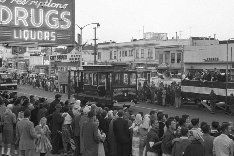 Black and white photograph from 1959 showing crowds of people lining Geary Boulevard at 18th Ave for a parade. Muni motorized cable car 62 is traveling down the street in front of a bandstand with announcers and a long line of storefronts.