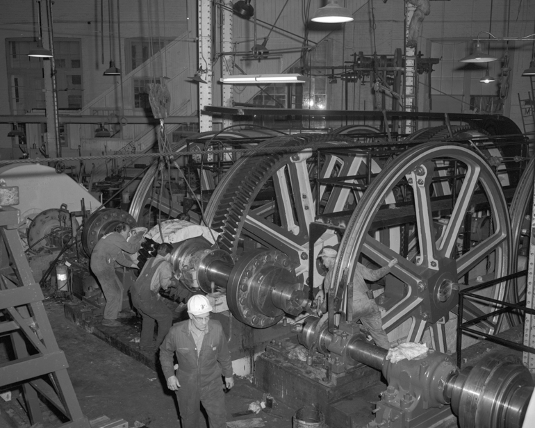 black and white photo showing workers lowering a large geared driveshaft into place next to huge pulleys used to pull cable car cables