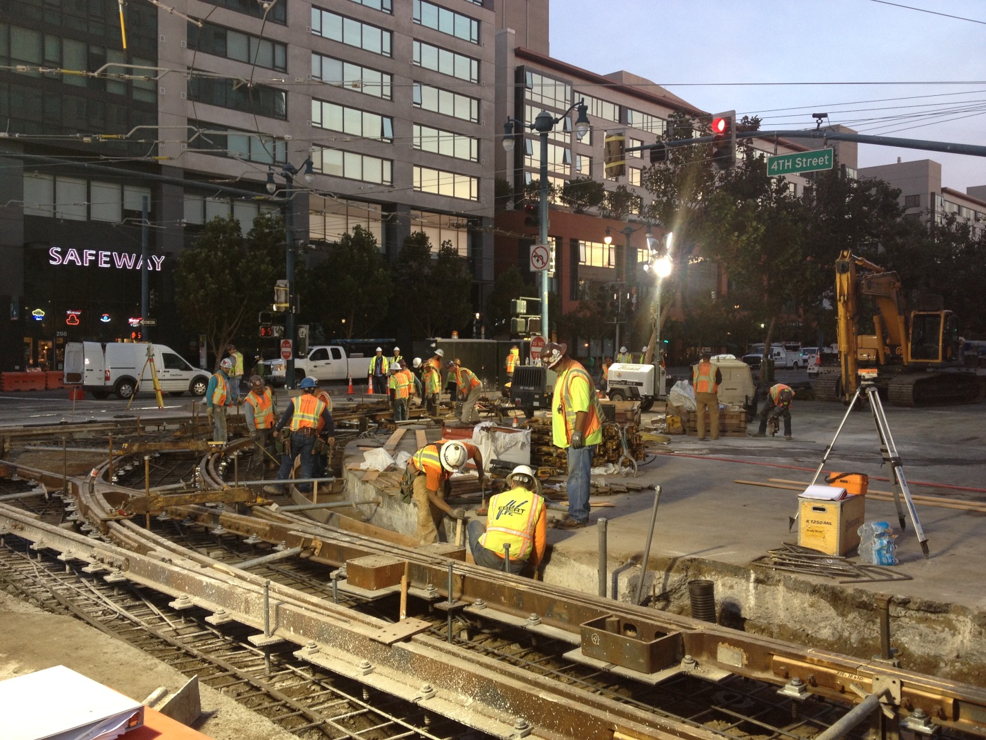 Crews in orange safety vests and hard hats work in the 4th/King trackway under large lamps at sunset with "Safeway" sign in the background.