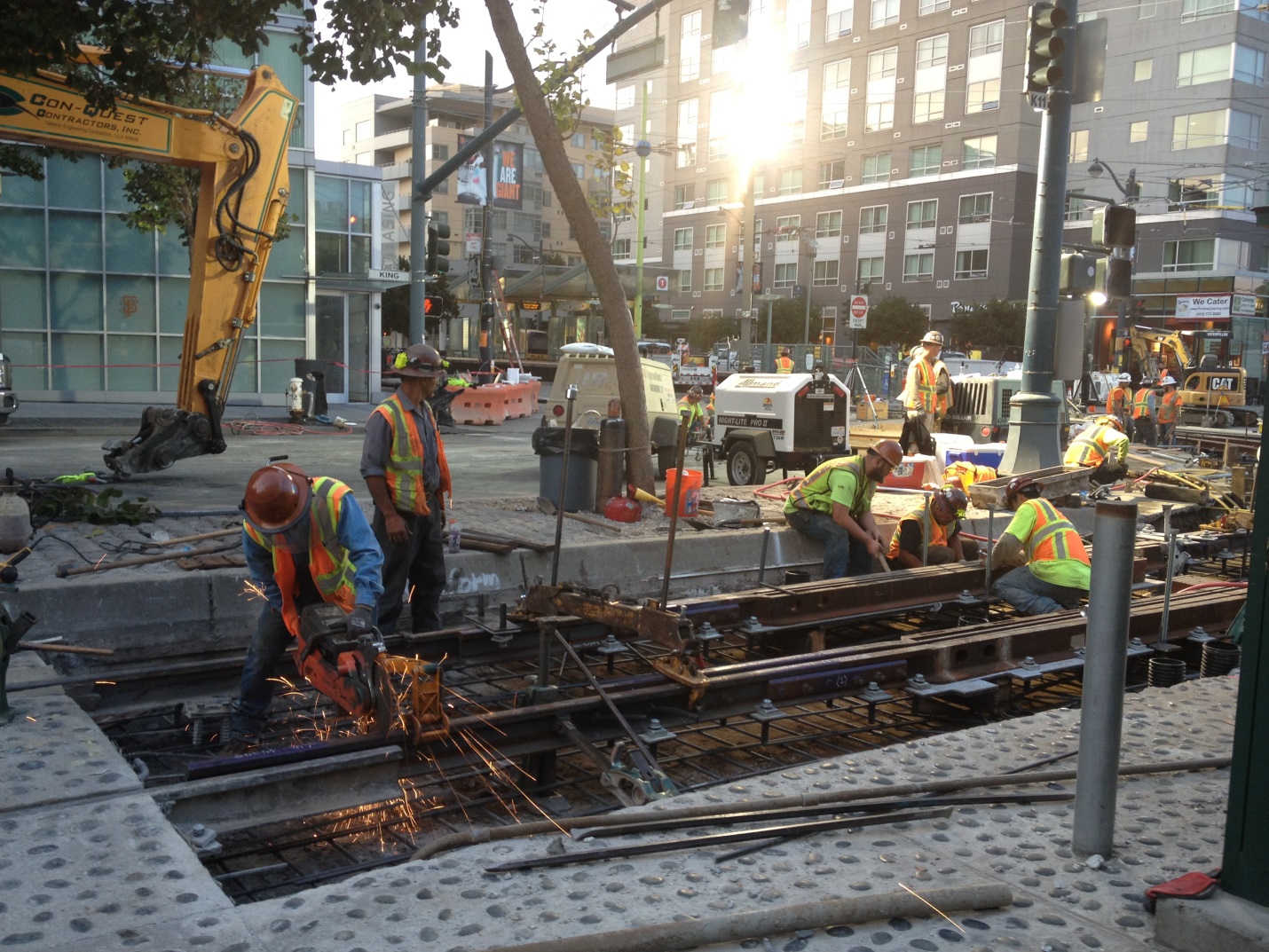 Crews in safety vests and hard hats work in pits along the tracks on King Street with the sun brightly reflected in the building behind them.