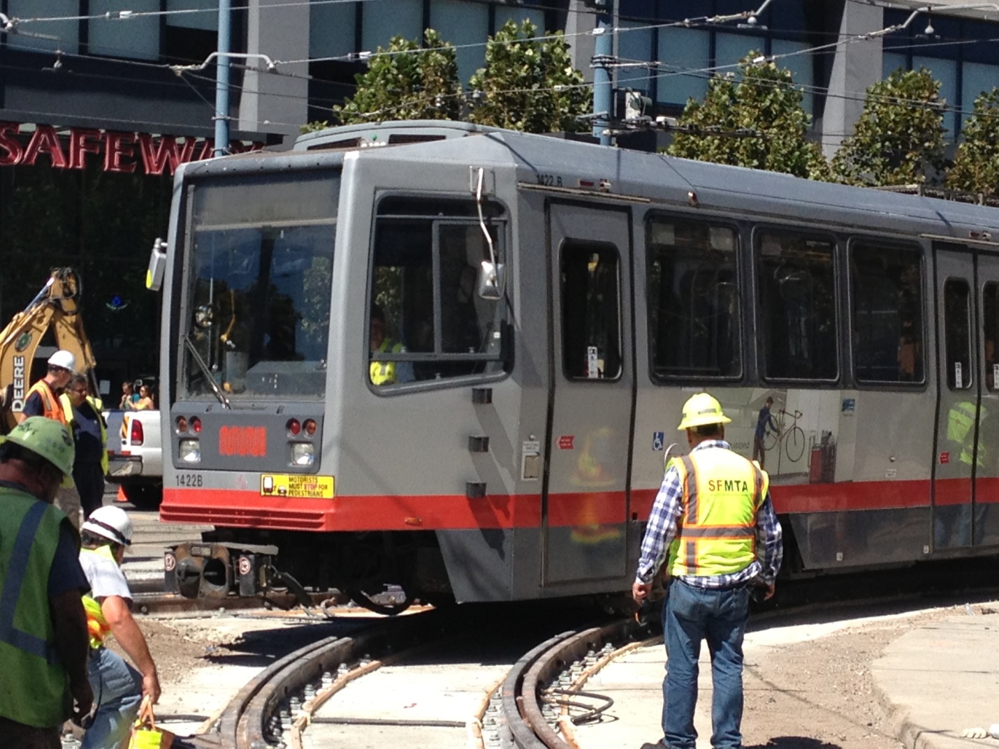 Gray and red Muni light rail train travels slowly through the construction area at 4th/King while crews and operations staff in safety vests and hard hats look on.