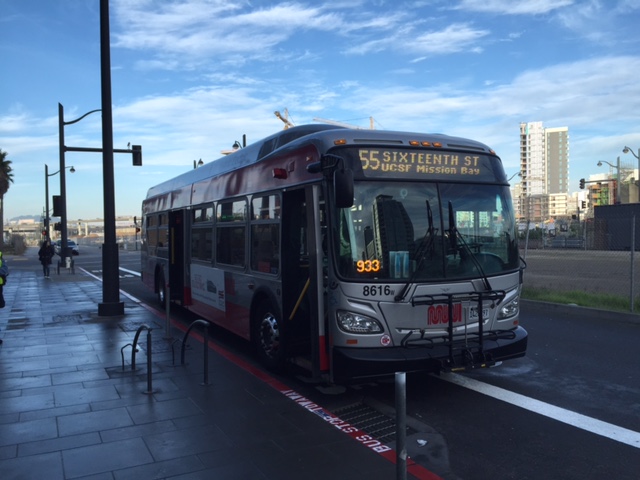 Muni hybrid "55 Sixteenth St, UCSF Mission Bay" bus sits at the curb with Mission Bay construction in the background.
