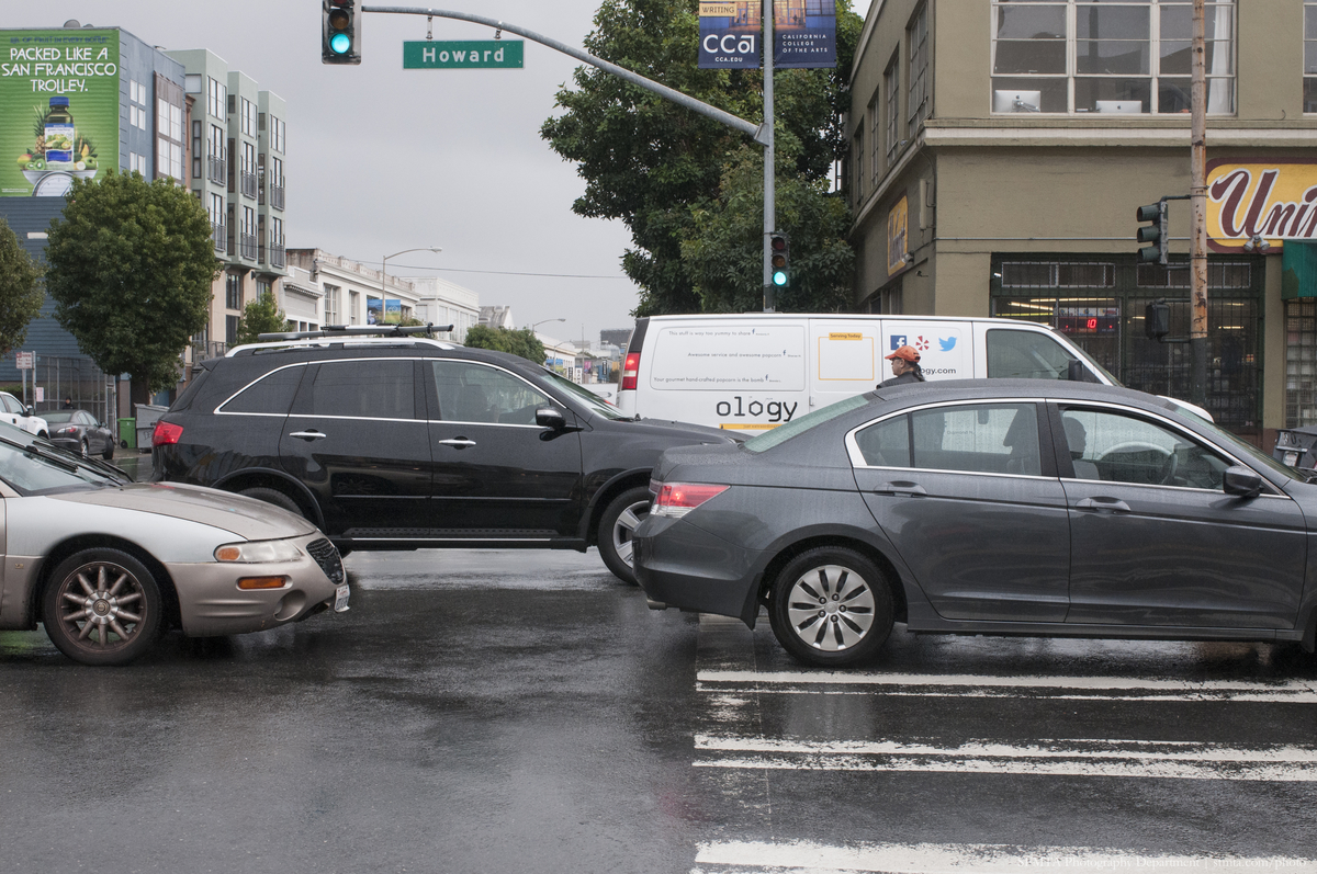 Traffic on a wet street in SoMa sitting in an intersection and over a crosswalk.