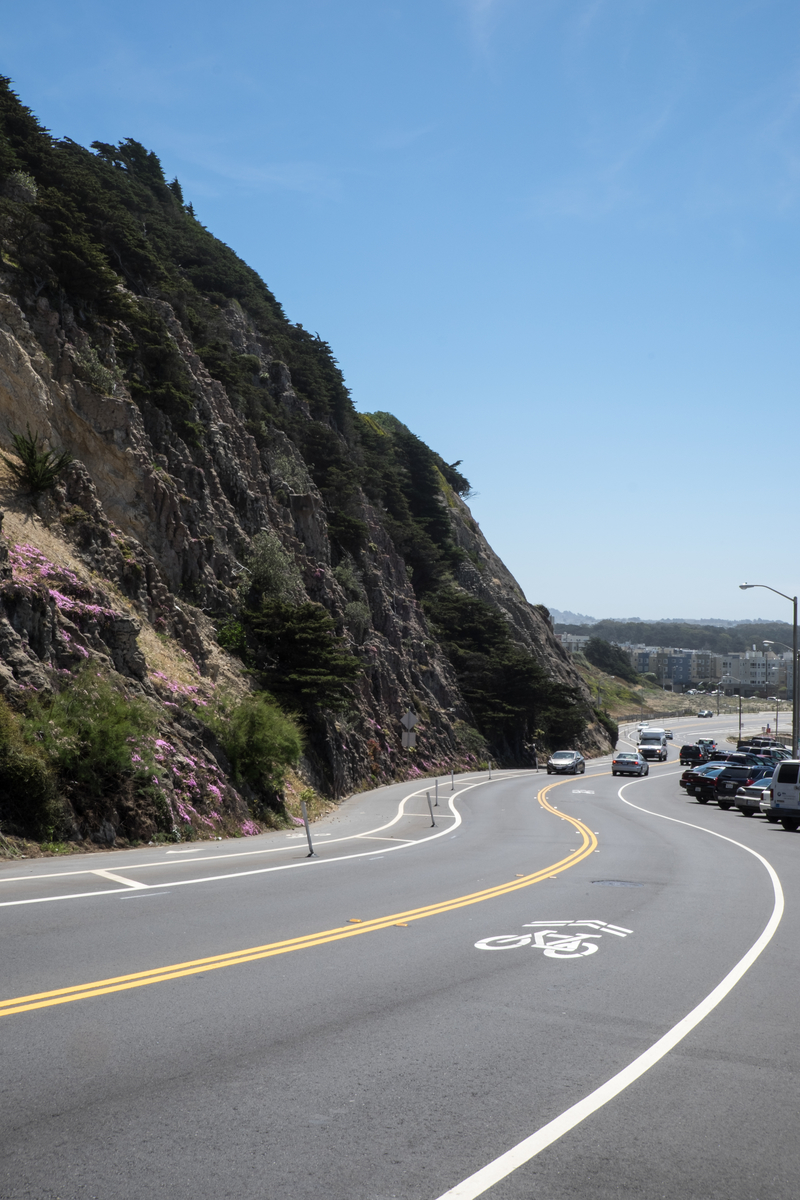 The curving road with a steep cliff rising on the left slopes down to Ocean Beach with a white road marking of two arrow and a bicycle.