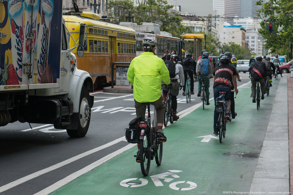Bicyclists in bright clothing and helmets cycle eastward on the bright green bike lane on Market Street with a bus, a streetcar and a delivery truck in the background.