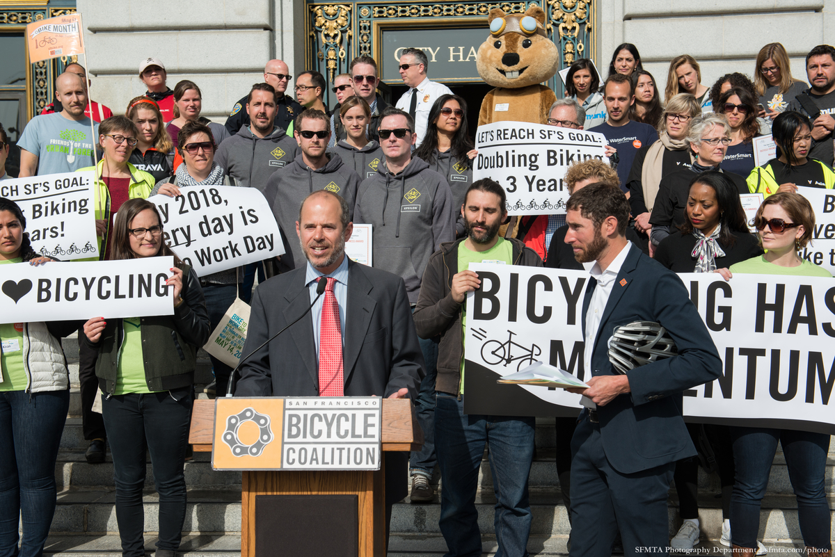 Ed Reiskin stands at a lecturne with San Francisco Bicycle Coalition Executive Director, Noah Budnick. Volunteers and participants stand with signs and banners on steps behind them.