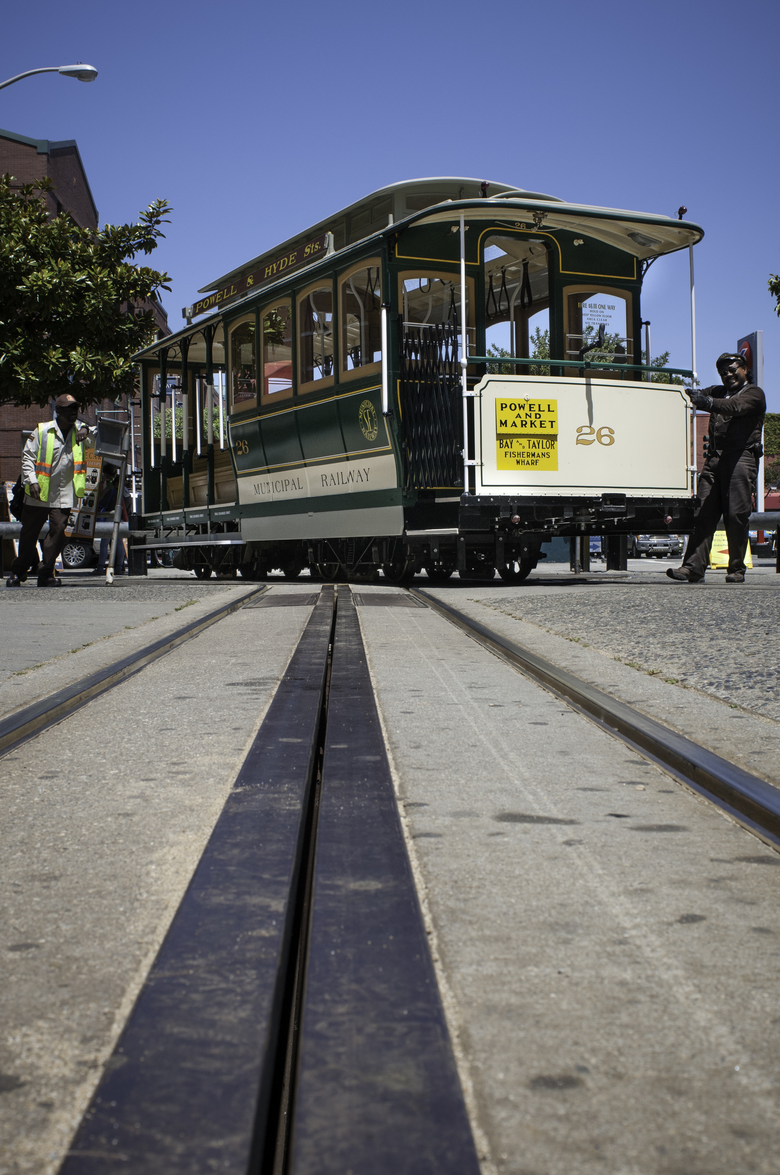 A cream and green cable car is being turned by the gripman and conductor in Muni uniforms. The low-angle shot captures the cable car tracks and slot in the foreground.